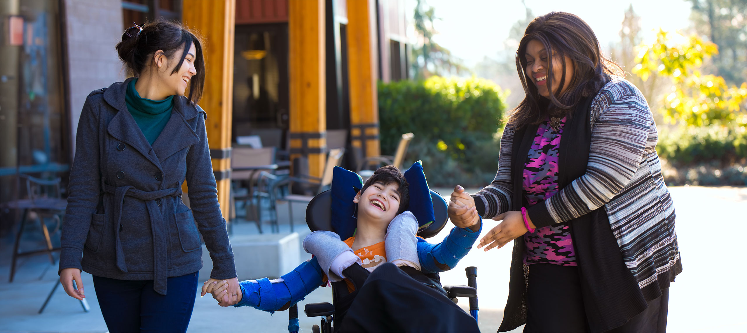 Two women who are WTC Support Coordinators smiling and supporting a joyful young person with a disability in a wheelchair—Coordinated Family Support in Riverside and San Bernardino Counties through WTC Support Services.